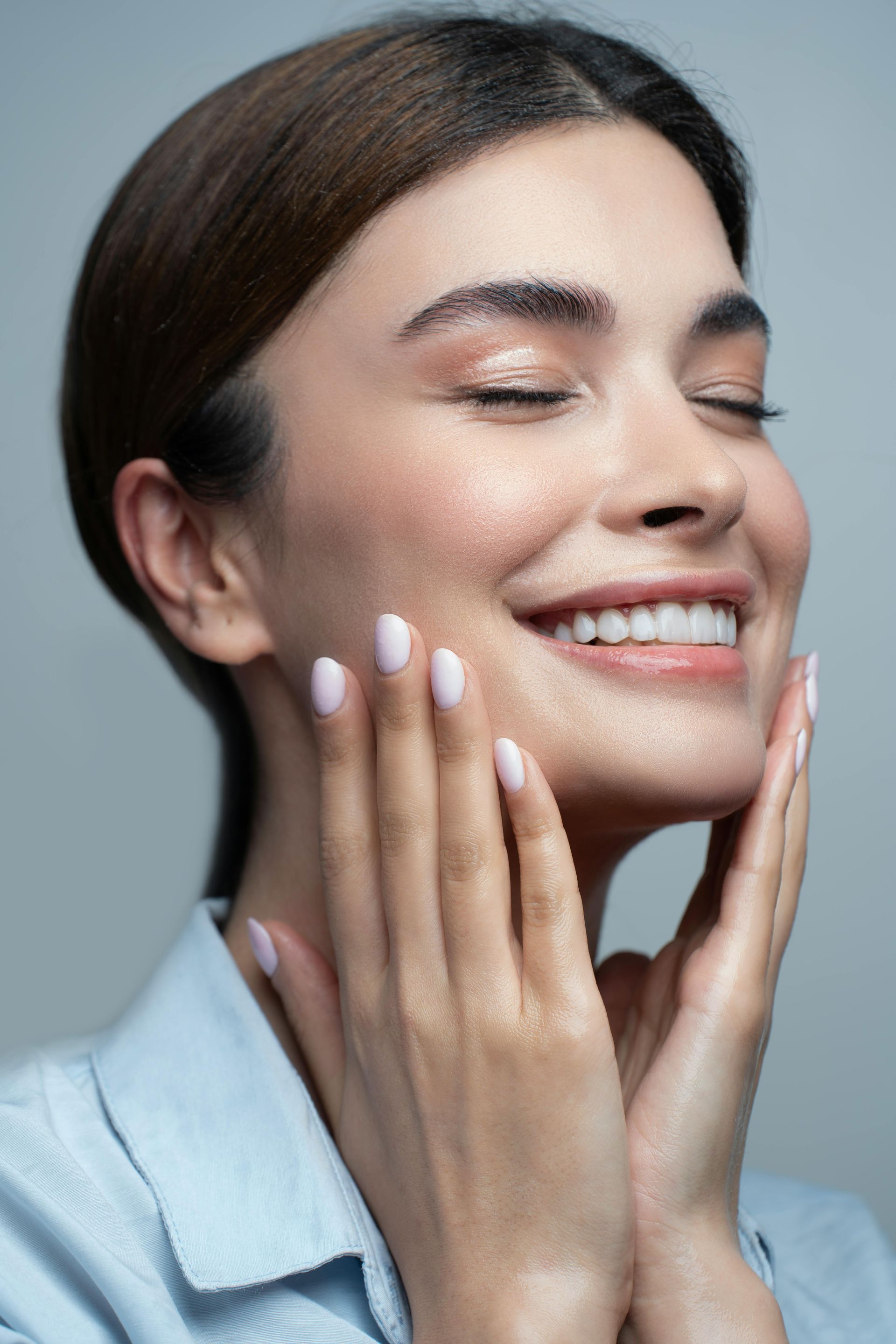 Woman with closed eyes smiling, touching face with hands, light blue shirt.
