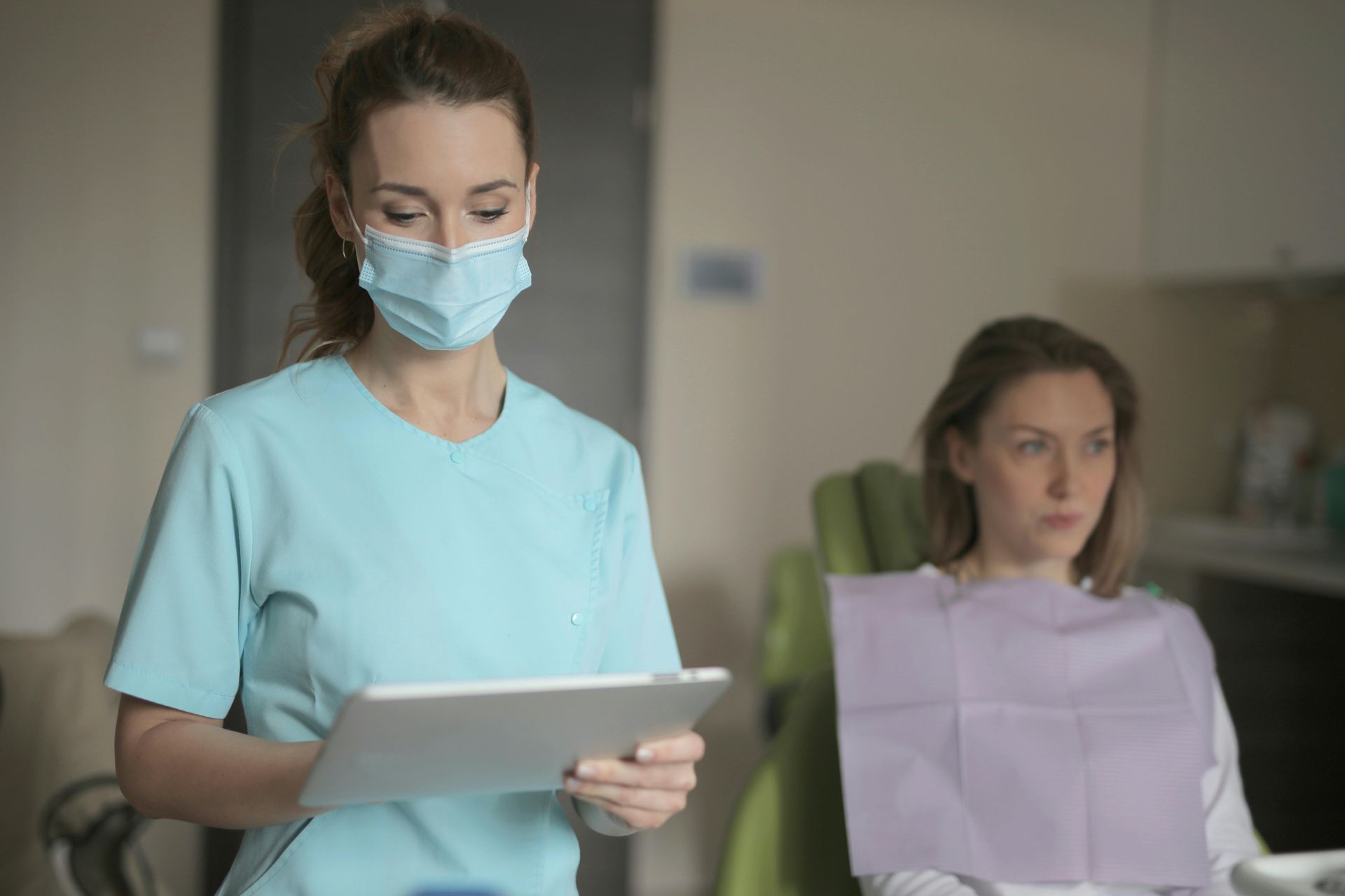 Dentist in scrubs and mask with tablet, talking to a patient in the dental chair.