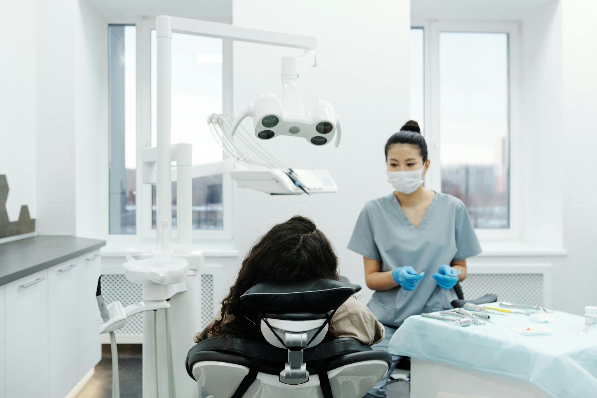 A dental assistant in scrubs and mask prepares tools next to a patient in a dentist's chair.