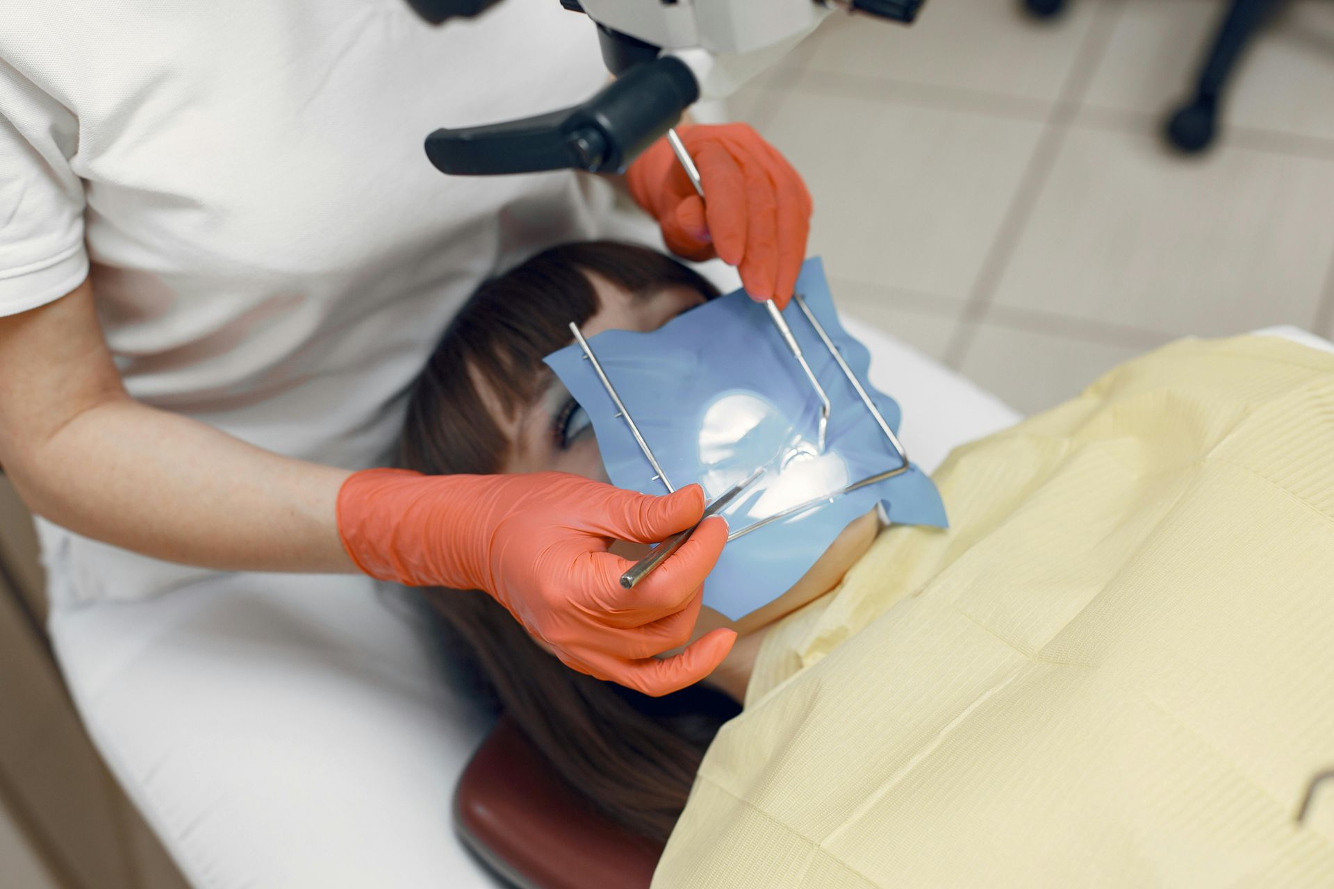 Dentist using a dental dam on a patient, orange gloves, dental microscope overhead.