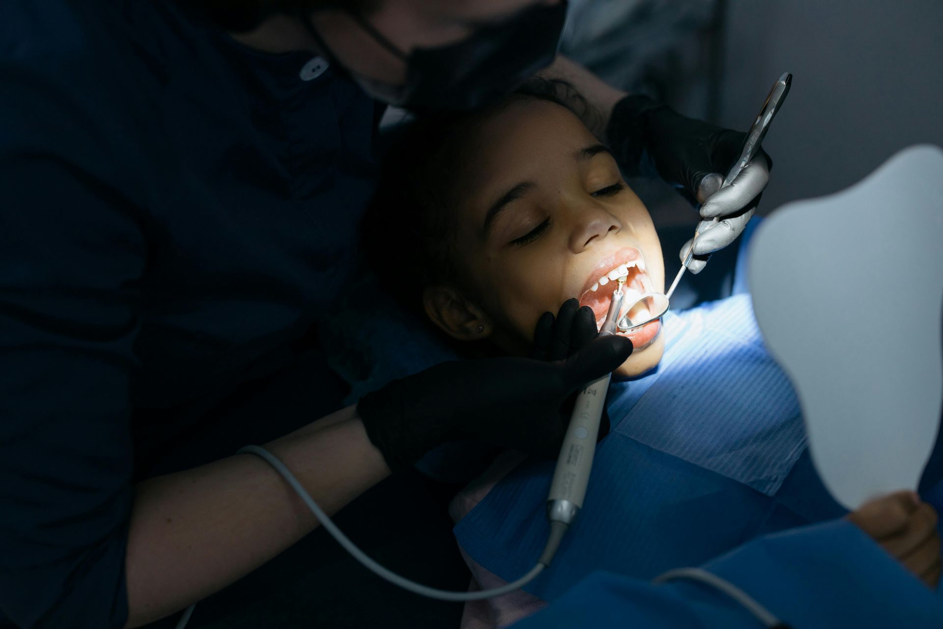 Child in dental chair, mouth open while dentist works with tools; bright light, blue drape.