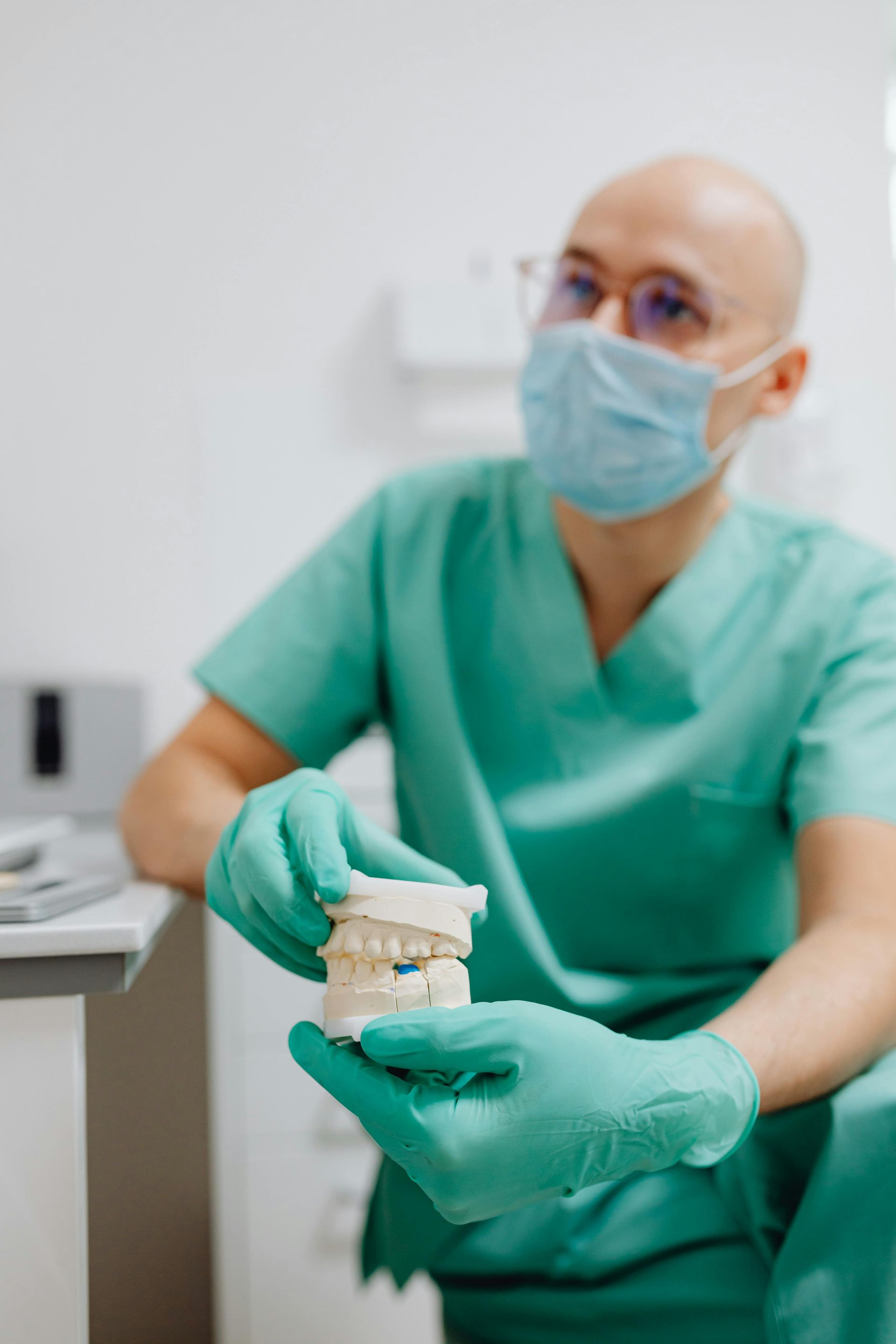 Dentist in teal scrubs and mask holding a dental mold, in an office.