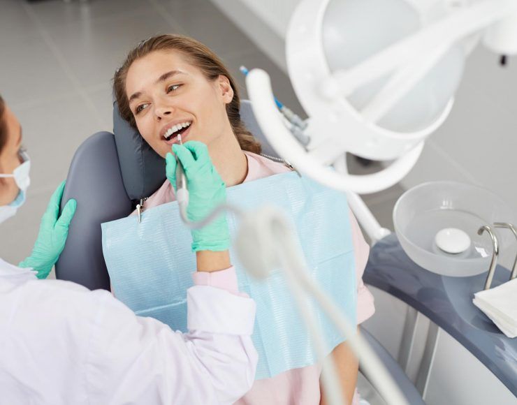 Dentist examining a patient's teeth. The patient smiles, blue bib, green gloves. Clinic setting.