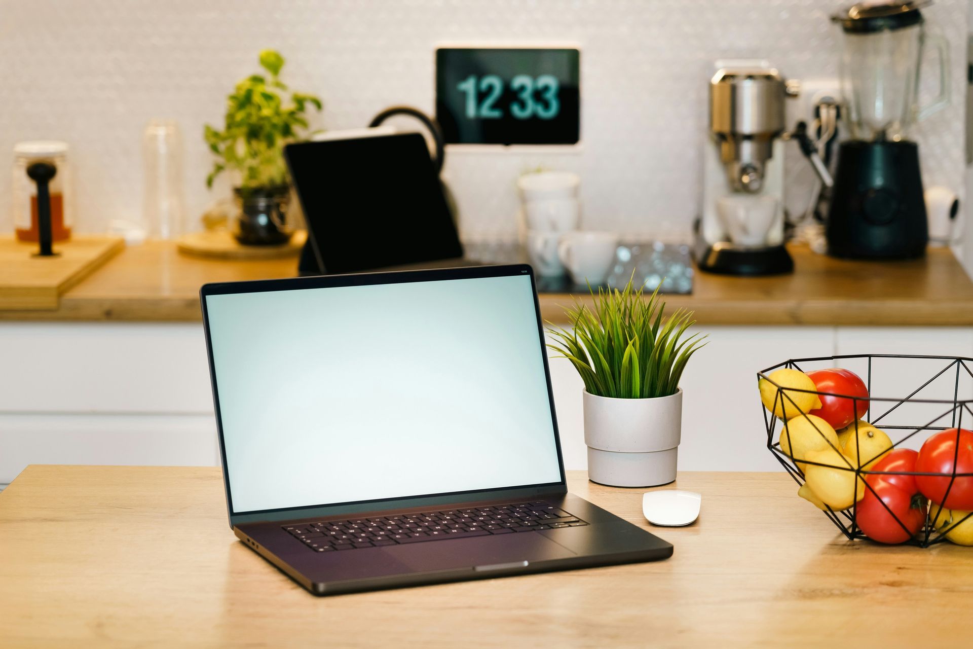 Laptop on a wooden counter with a mouse, plant, and fruit basket. Kitchen in background.
