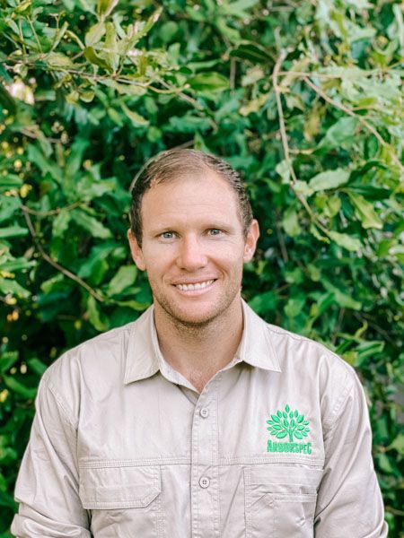 A man in a tan shirt is smiling in front of a tree.