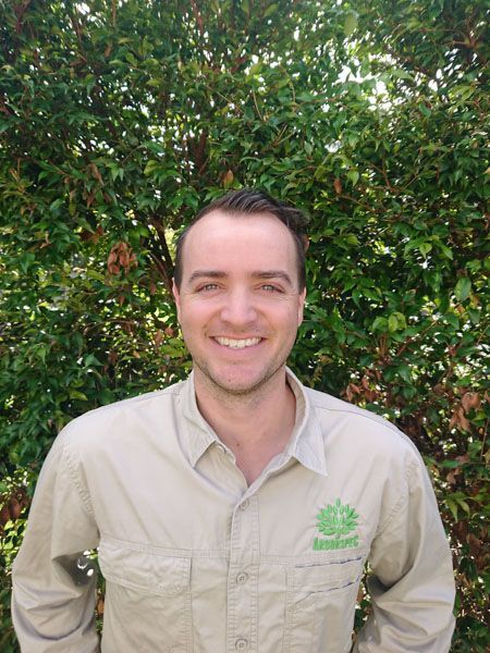 A man in a tan shirt is smiling in front of a bush.