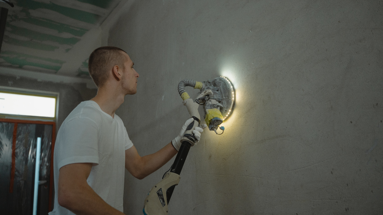 Man sanding a wall with a drywall sander, illuminated by a bright light. Interior shot.