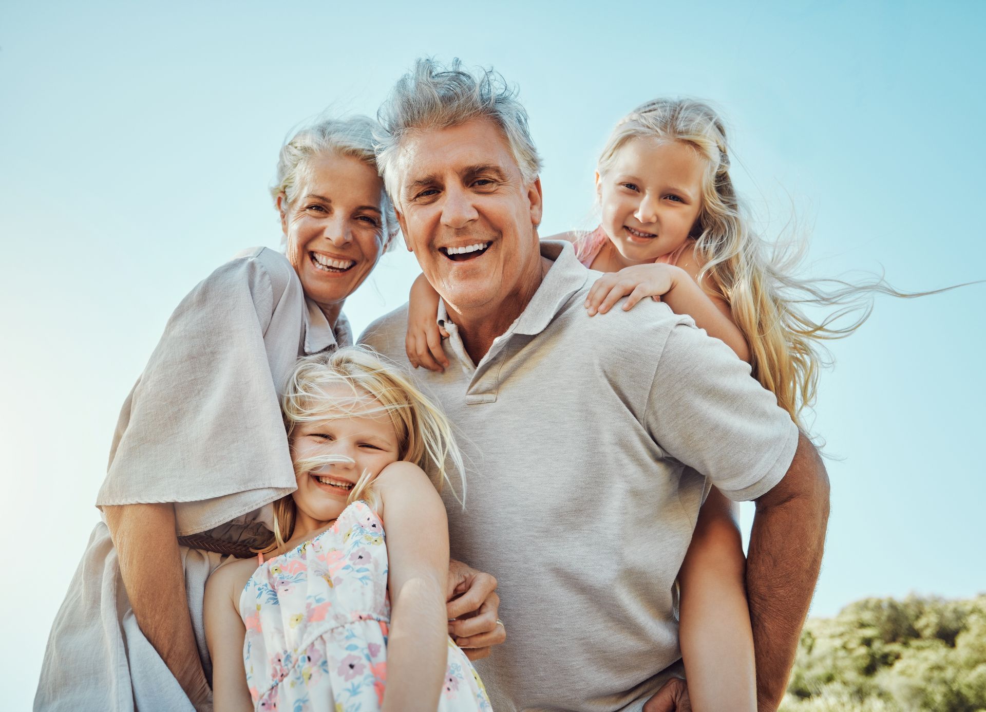Two adults and two children embrace outdoors against a clear blue sky, all smiling happily.