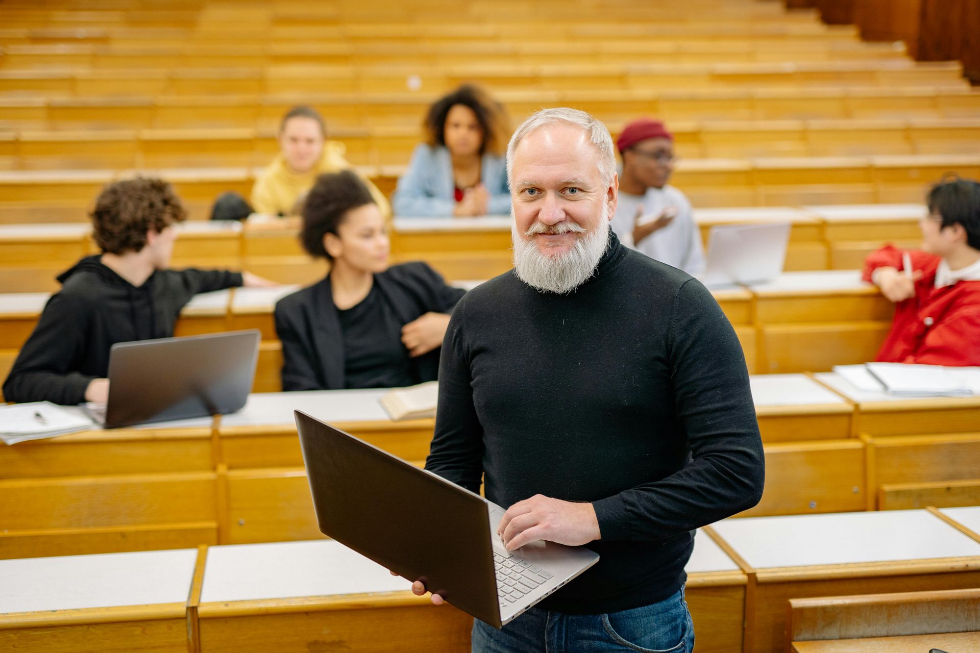 A man with a beard is holding a laptop in a lecture hall.