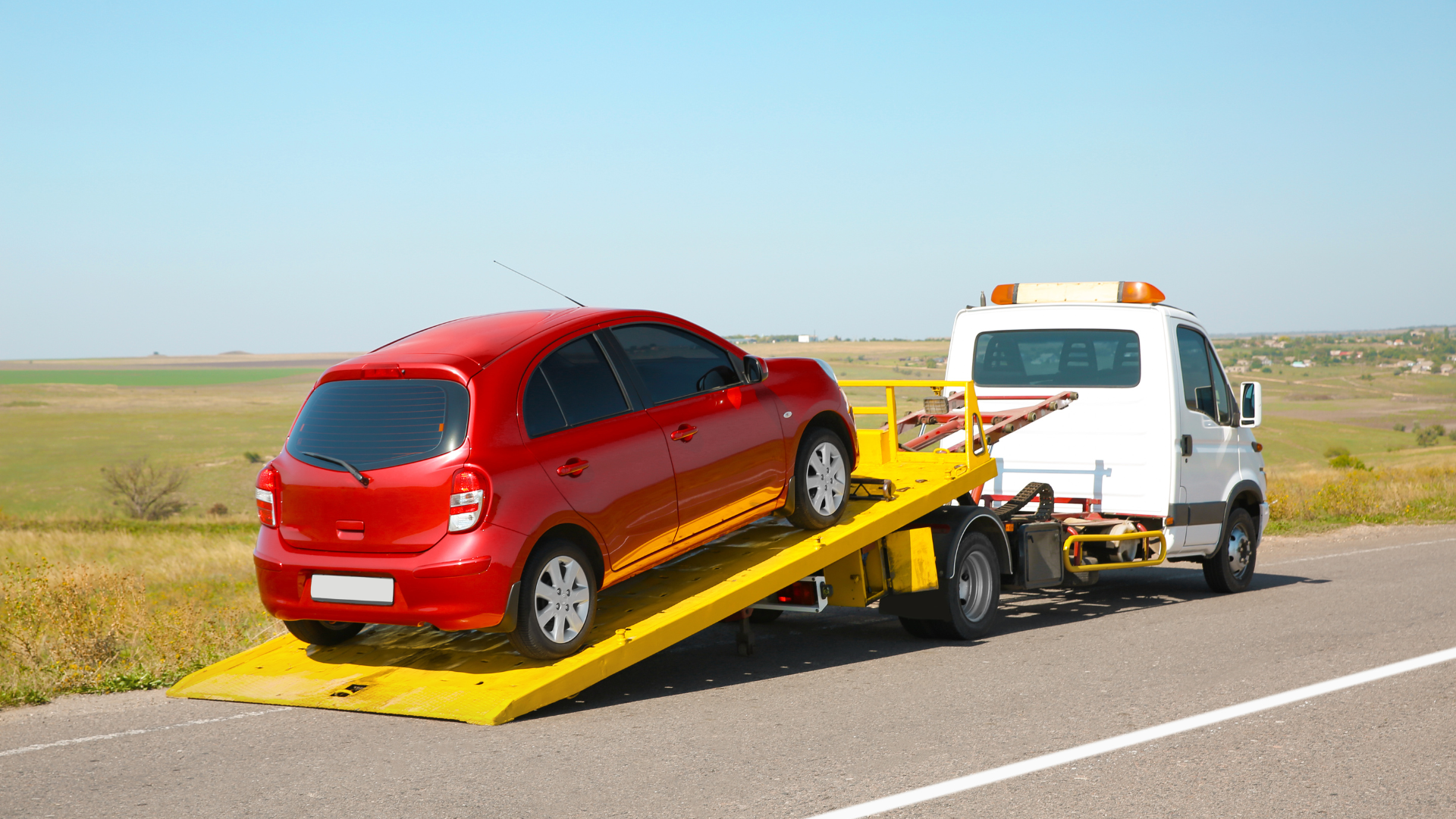 A red car is being towed by a tow truck on the side of the road.