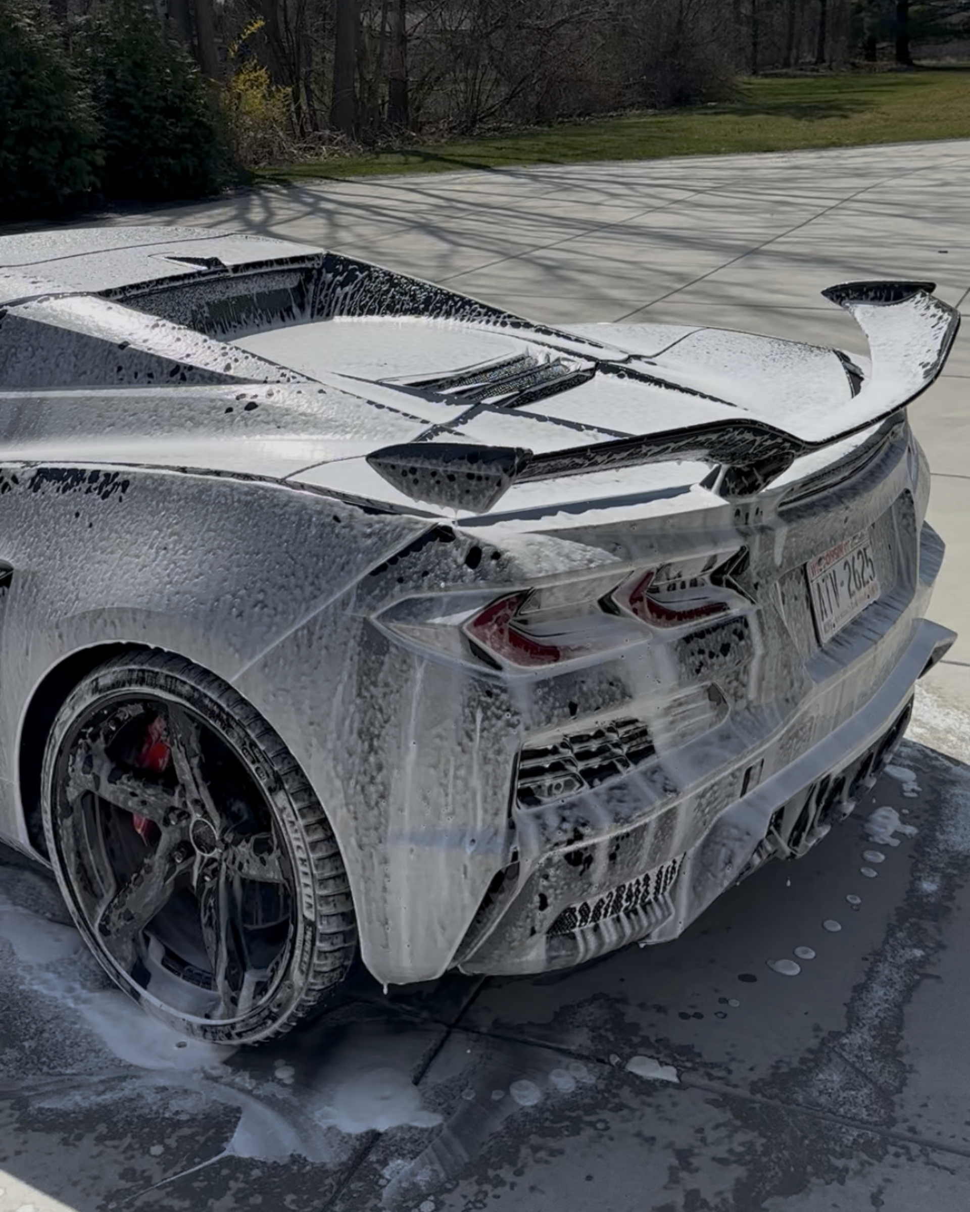 White sports car rear, covered in mud or snow, parked outdoors near leafless trees.