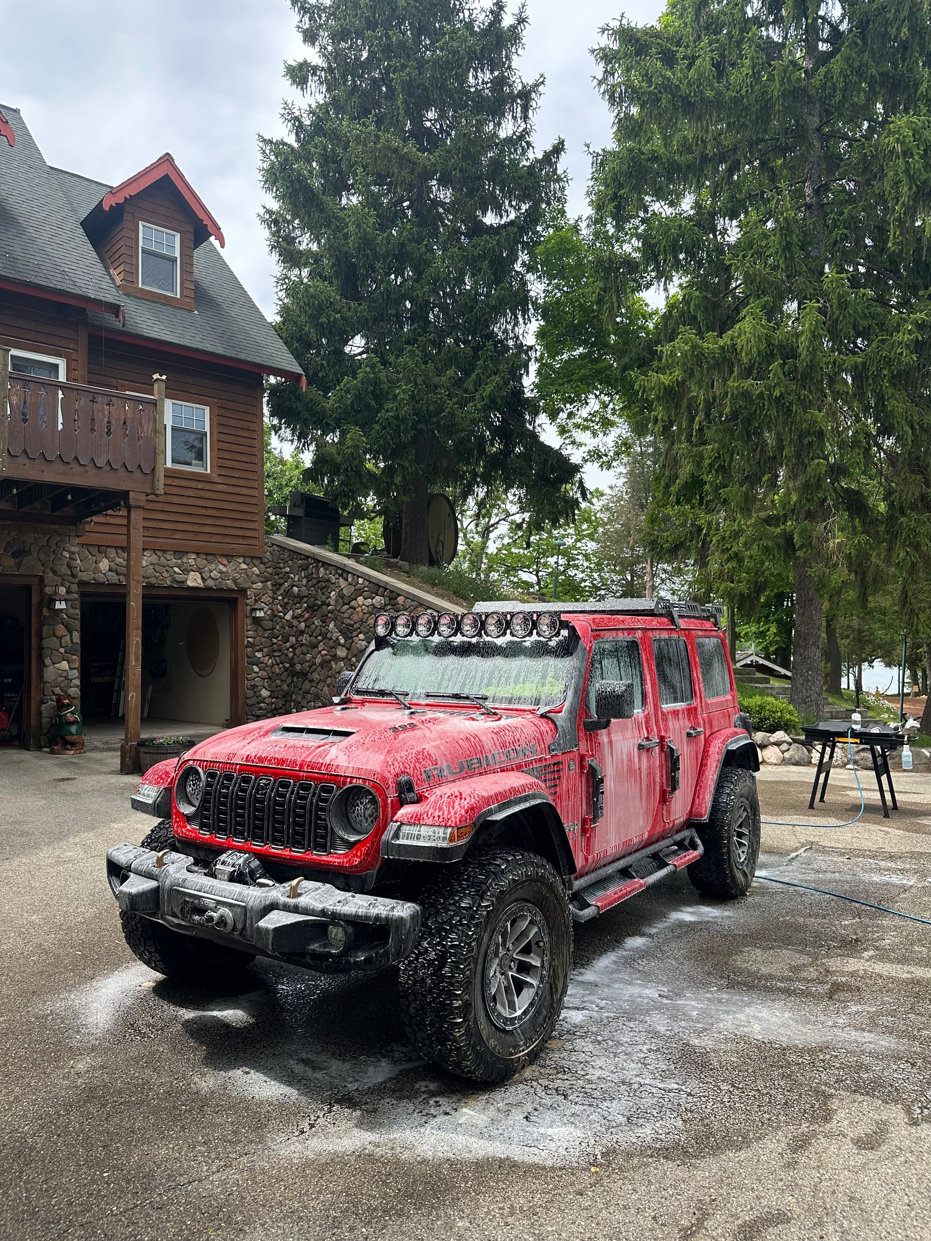 Red Jeep Wrangler parked on a wet gravel driveway beside a rustic house and trees.