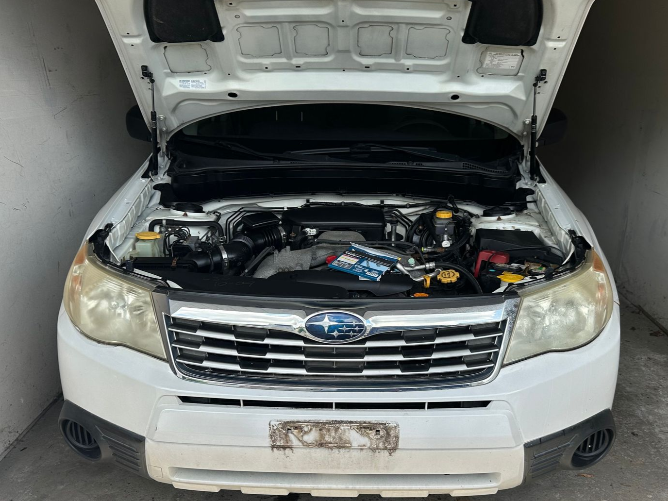 White Subaru sedan with hood open, showing the engine bay in a garage-like setting