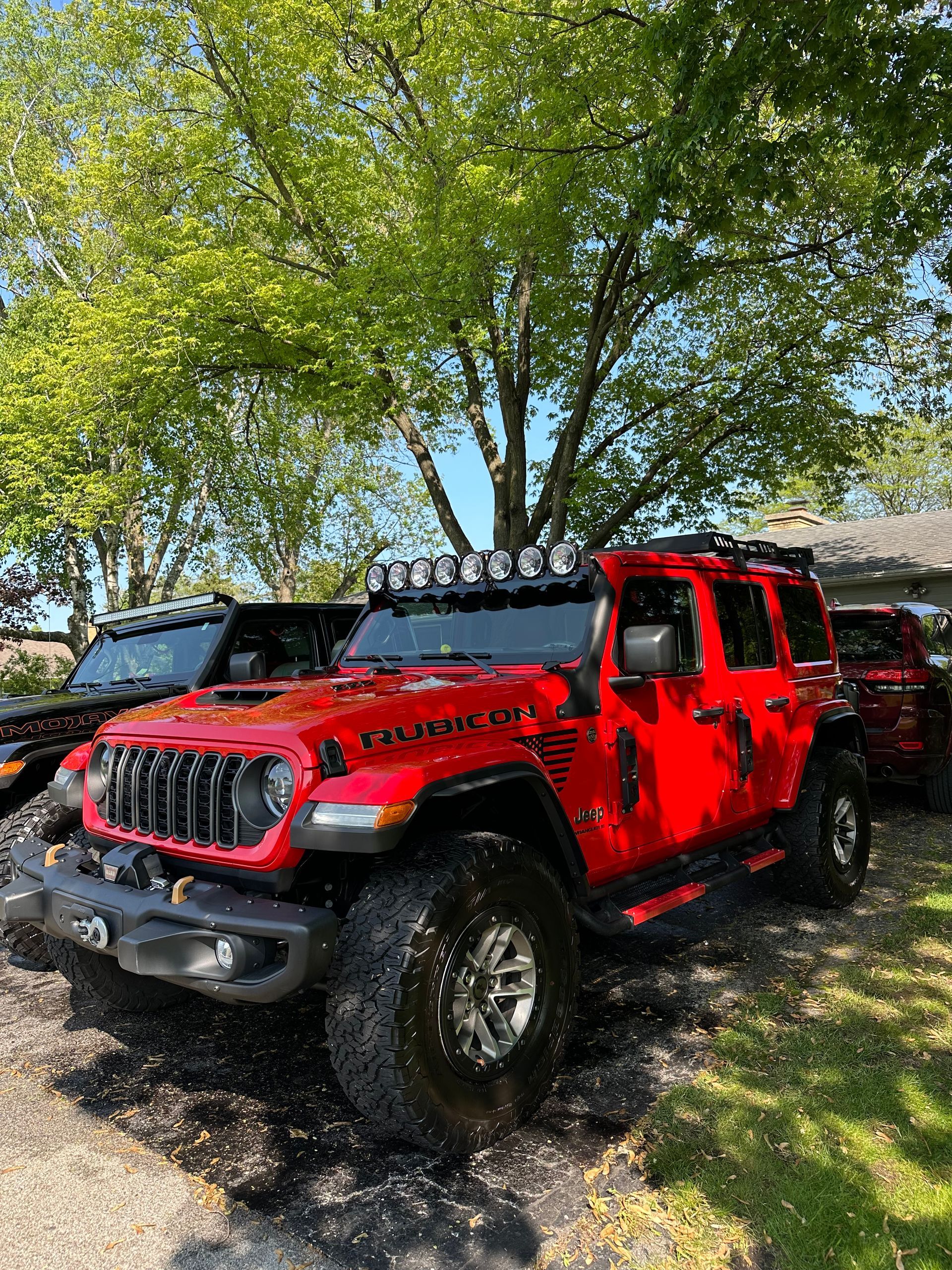 Red Jeep parked under trees on a sunny dirt lot