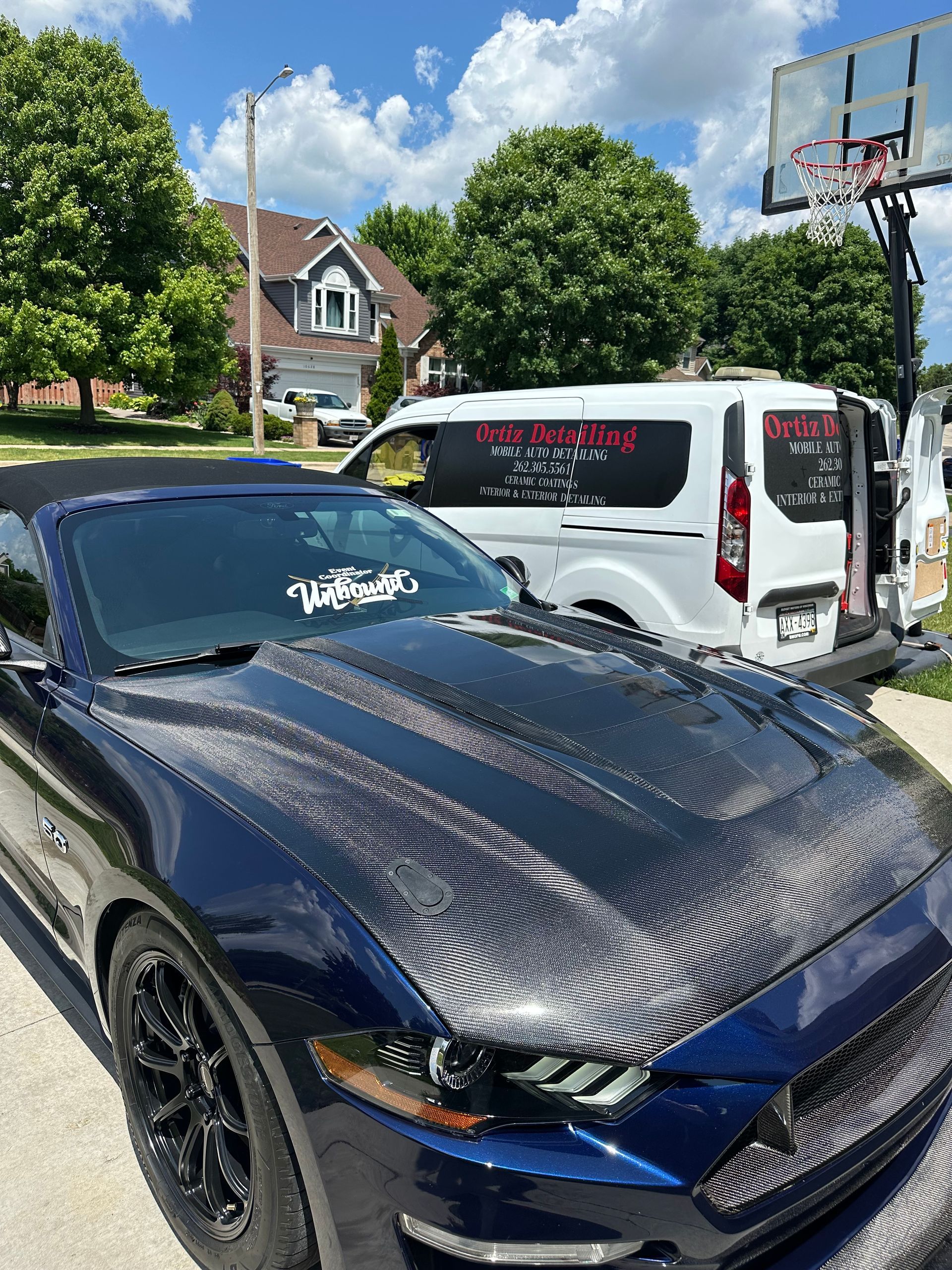 Black sports car with carbon-fiber hood parked beside a white van on a sunny suburban street.