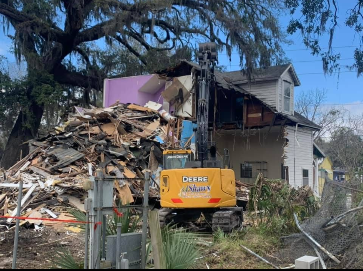 Yellow excavator demolishing a house, debris pile, trees, blue sky.