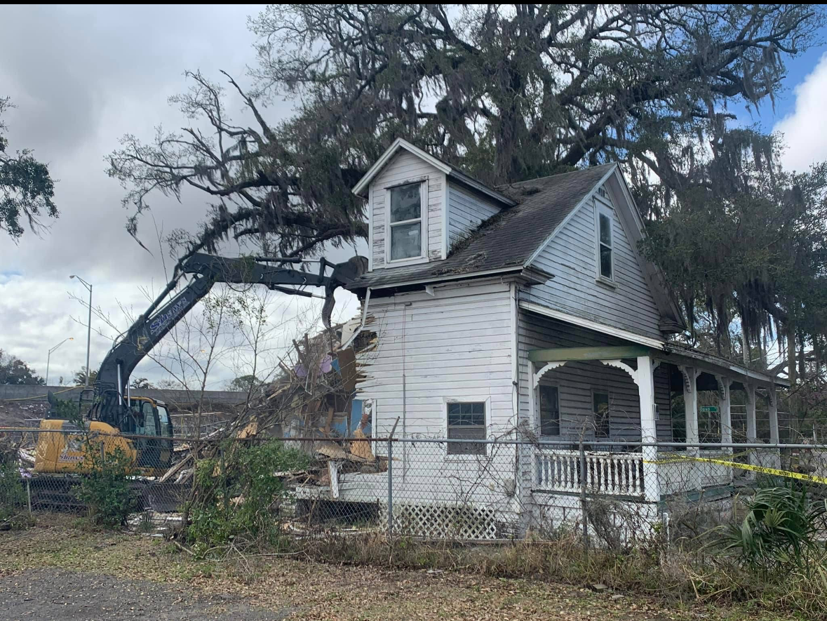 An excavator demolishes a weathered, two-story house with a porch and overgrown trees.