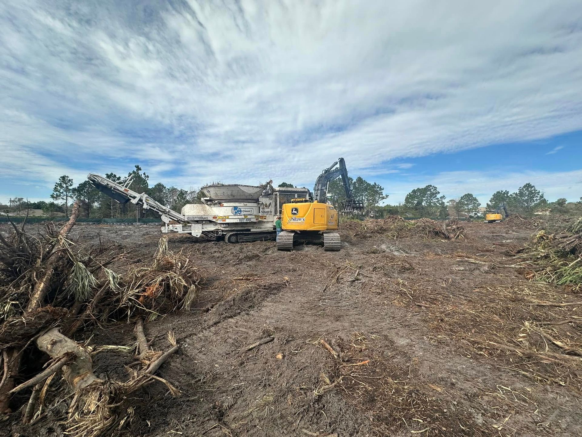 Heavy machinery clearing land, wood chipper and excavator visible. Cloudy sky overhead.