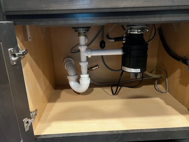Under-sink plumbing with garbage disposal in a kitchen cabinet. White pipes, dark disposal.
