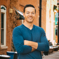 A man in a blue shirt is standing with his arms crossed in front of a brick building.