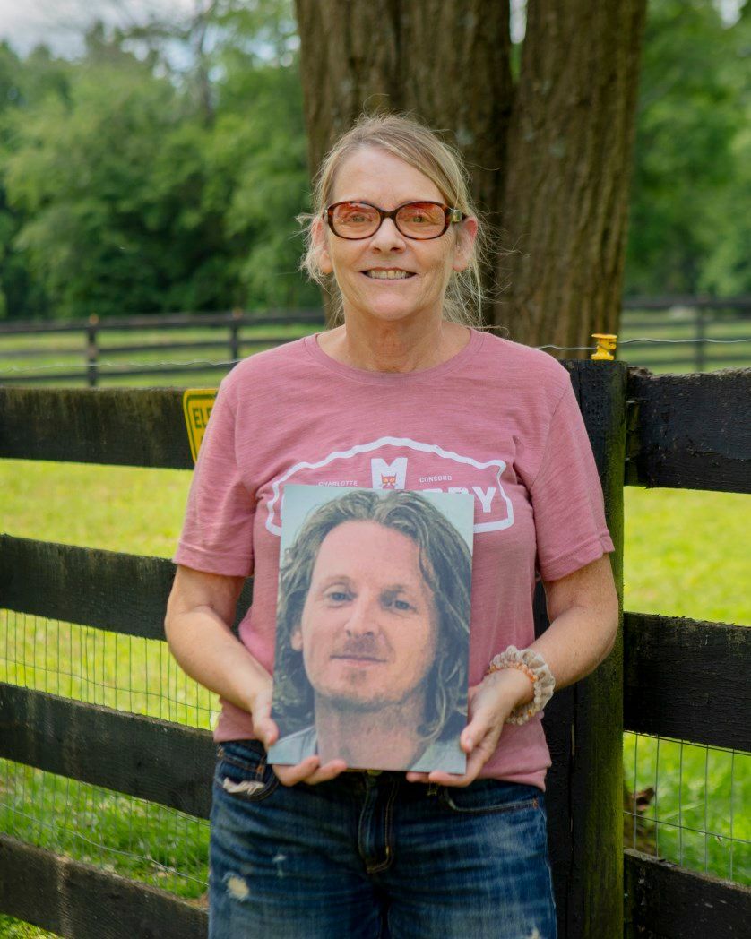 A woman in a pink shirt is holding a picture of a man with long hair.