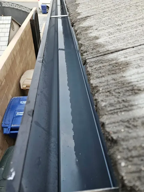 Black rain gutter filled with water, next to a concrete roof edge, overlooking a blue trash bin.