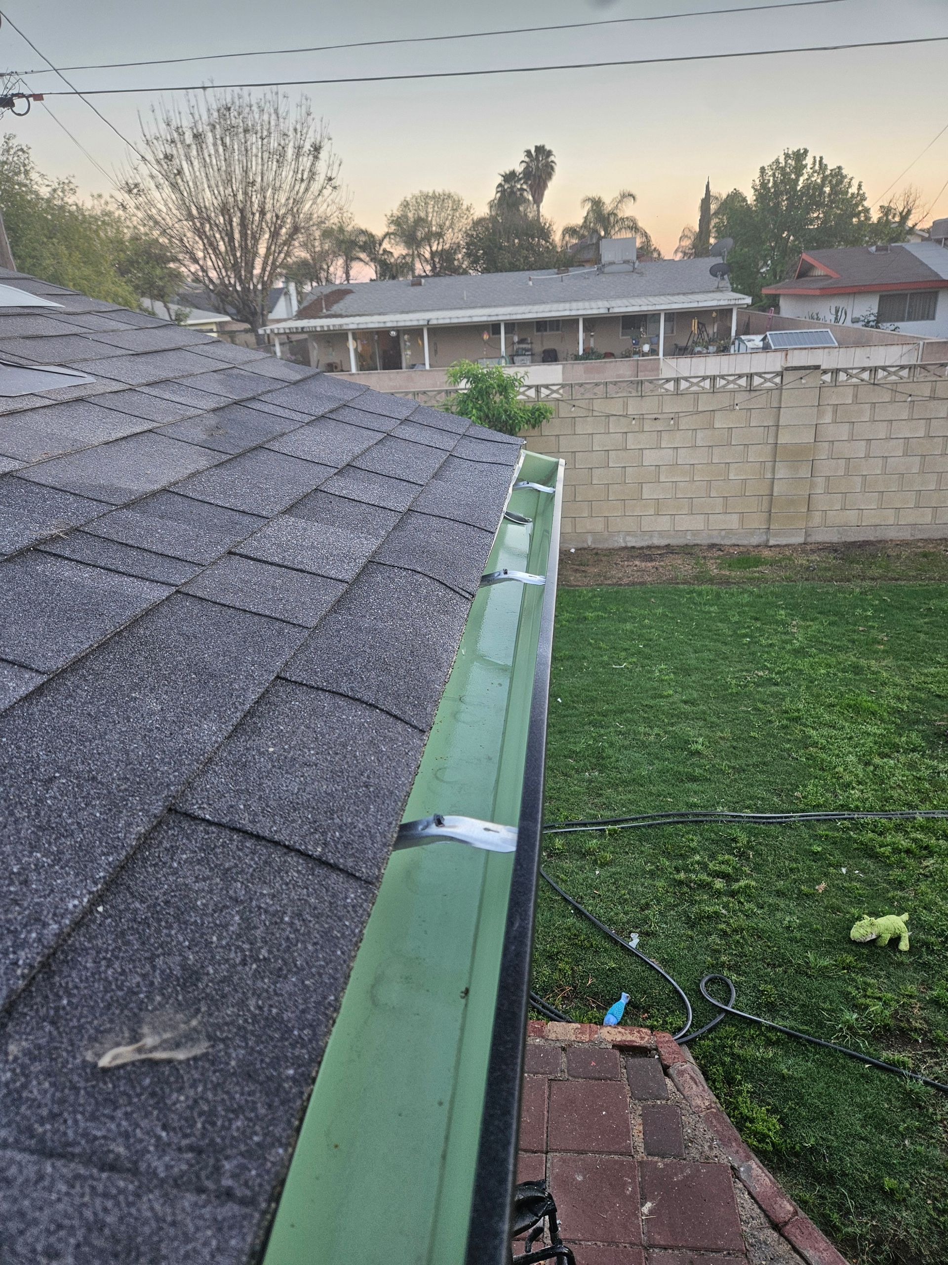 A view looking down along a green gutter attached to a dark shingled roof, with a yard and suburban homes in the background.