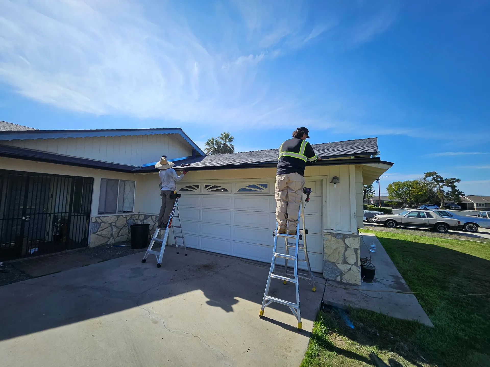 Two workers on ladders, repairing a roof on a sunny day. House has a garage door and stone accents.