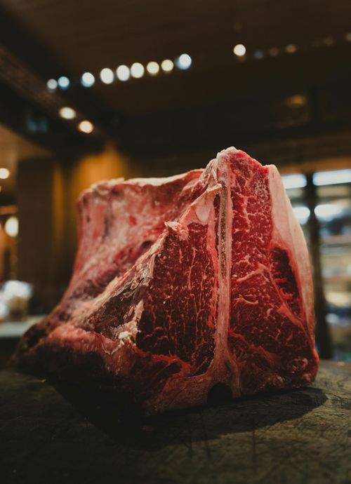 Large, raw T-bone steak on a dark surface, red meat with white marbling and bone visible.