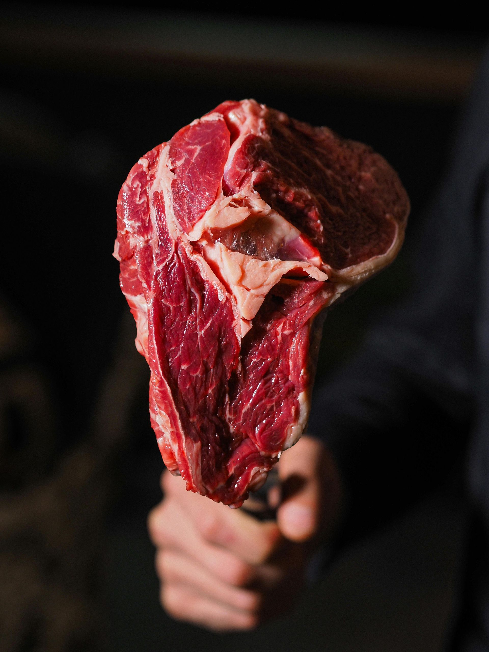 A raw, thick-cut steak, held up by a hand against a dark background, showing red meat and white fat.