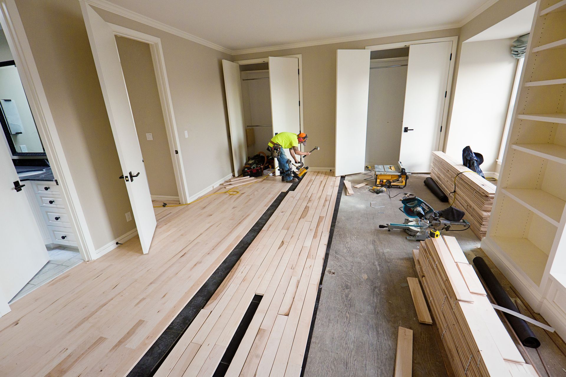 A room under construction with a worker installing hardwood flooring. The room has built-in shelves, closets, and a partially installed floor.