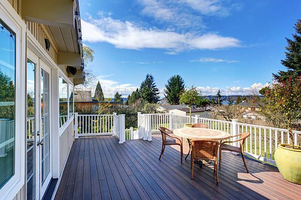Wooden deck with white railing, a table and chairs, and a view of trees and houses under a blue sky.