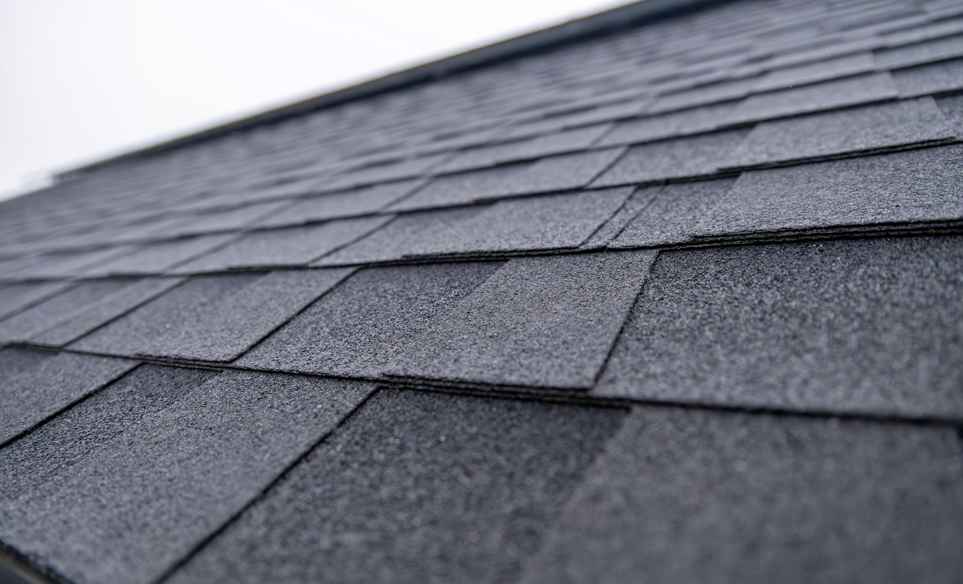 A close-up, angled view of dark grey asphalt roof shingles overlapping in a repeating pattern.