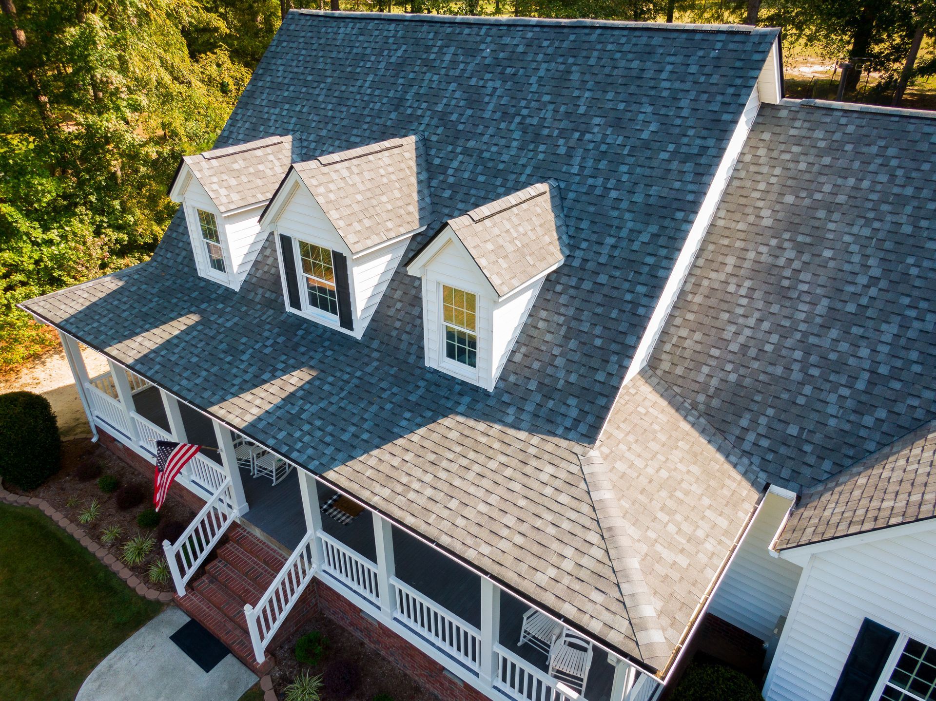 Aerial view of a white house with a multi-gabled dark gray shingled roof and a front porch, surrounded by green trees.