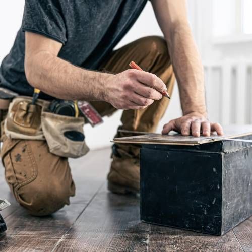 A man is kneeling on the floor measuring a piece of wood.