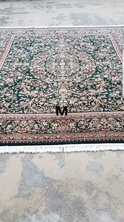 A dark green rectangular area rug with an intricate floral pattern, a central medallion, and fringed white edges.