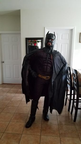 A person in a full Batman costume with a black cape, mask, and utility belt, standing in an indoor home setting.