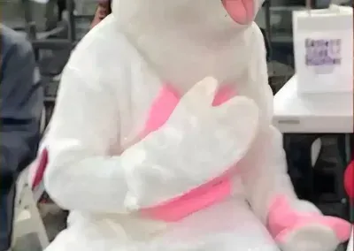 A mascot in a white and pink rabbit costume sits in a room, holding a hand over its chest.