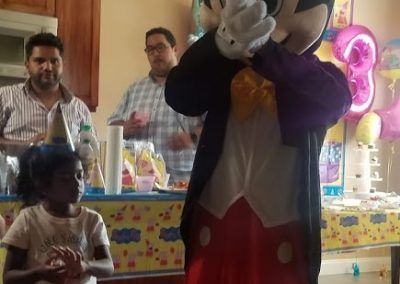 A person in a Mickey Mouse costume stands at a party with a young child and two adults in the background.