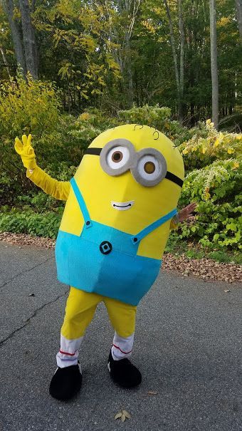 A person wearing a homemade Minion costume waves while standing on an asphalt path in a park with autumn foliage.