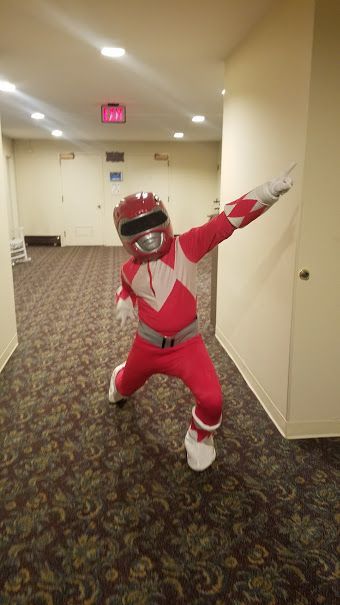 A person in a red Power Ranger costume posing with one arm extended in a hotel-style hallway with patterned carpet.