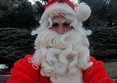 A person wearing a Santa Claus costume with a white wig, long curly beard, and red hat, posing for a selfie outdoors.