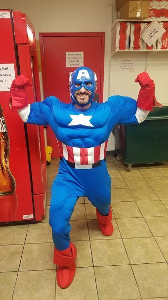 A person wearing a Captain America costume stands in a room with a red door, flexing their arms while smiling.