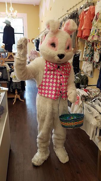 A mascot in a white rabbit costume, wearing a pink and white checkered vest, waves while holding an Easter basket.