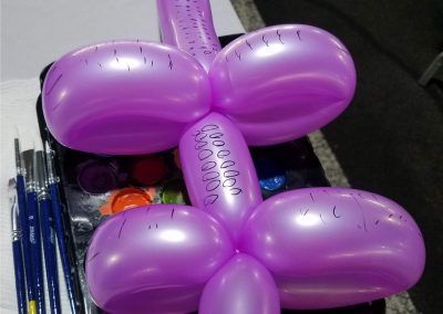 A purple balloon flower shape with handwritten text on it, resting on a paint palette with several art brushes nearby.