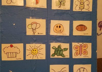 A grid of hand-drawn pictures on a blue board featuring items like a butterfly, cupcake, sun, turtle, and smiley face.