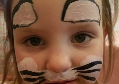 Close-up of a child with white face paint on their eyebrows and muzzle area, accented with black tiger-like whiskers.
