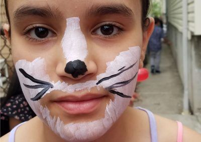 Close-up of a person wearing cat-themed face paint with white areas, a black nose, and whiskers on an outdoor patio.