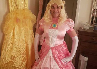 A smiling person wearing a pink Princess Peach costume with a blonde wig and white gloves, standing next to a yellow dress.
