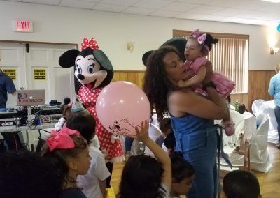 A woman holds a baby in a pink dress next to a Minnie Mouse mascot at an indoor party, with children nearby and a balloon.